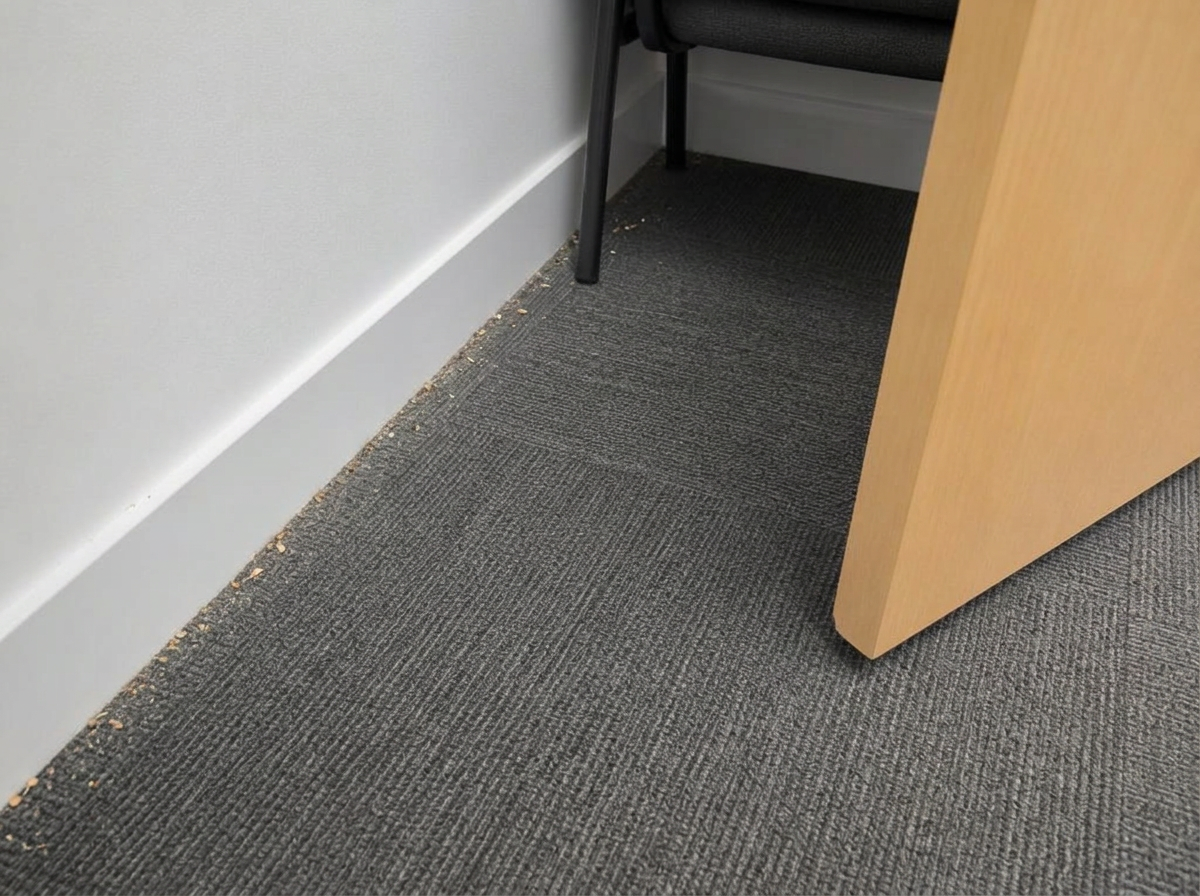 The same meeting room floor edge from the same angle: open floor clean, but a line of dust and crumbs still sits in the carpet-to-skirting edge