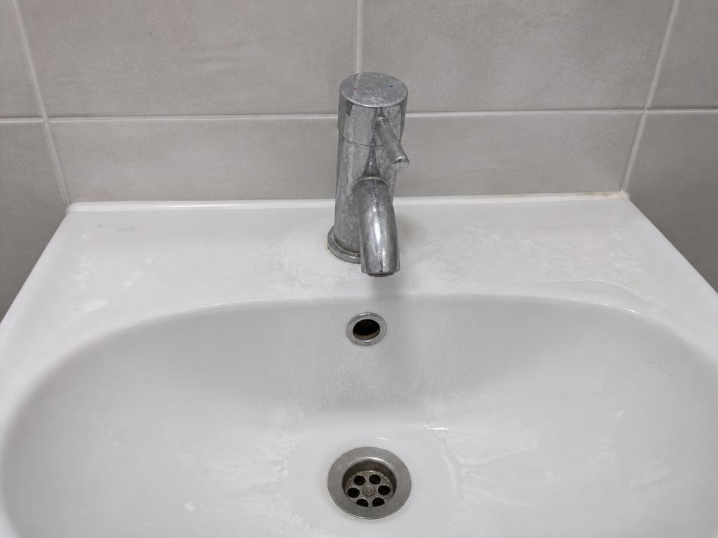 Washroom basin and chrome tap with dried water marks, residue and standing water around the tap base